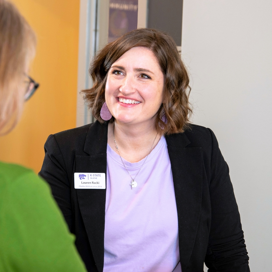 Lauren Racki smiles while speaking to a colleague. She is wearing a purple blouse and a black blazer.