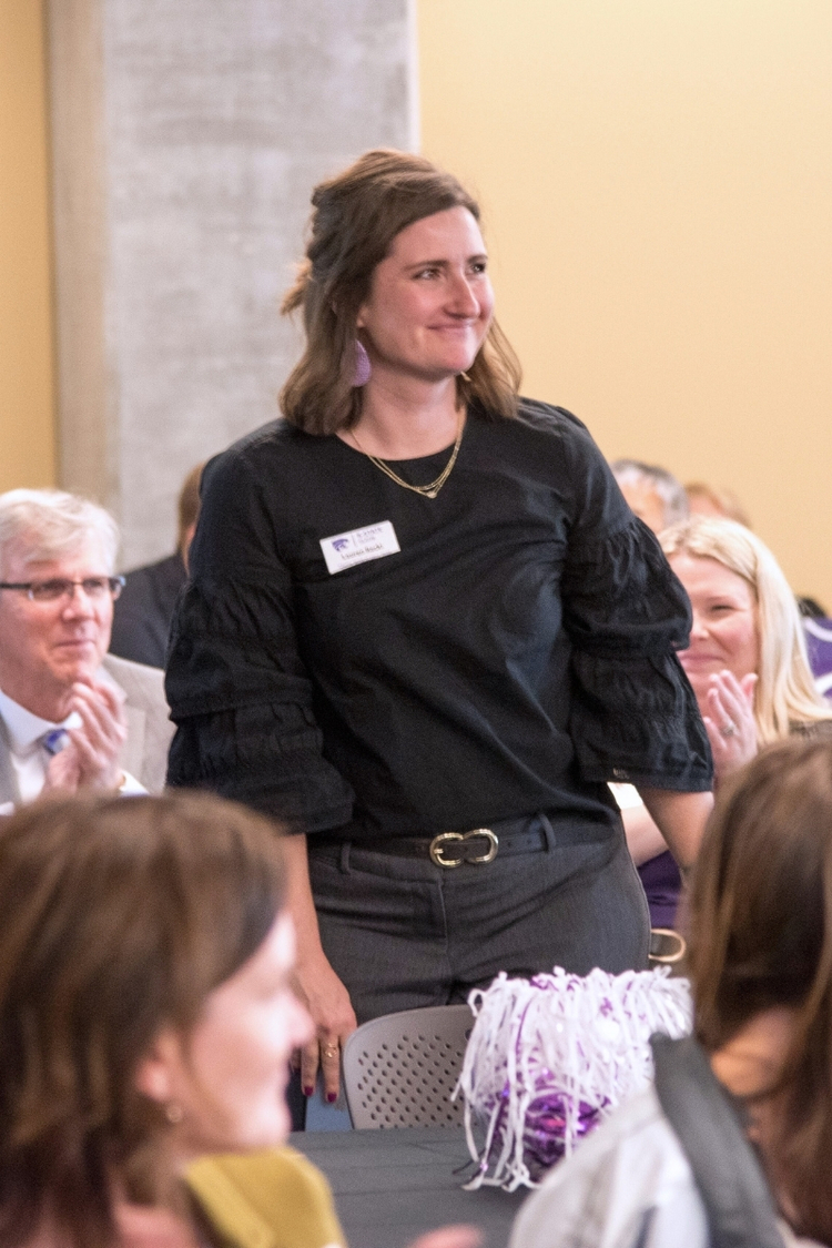 Lauren Racki stands smiling at a formal event while people seated around her applaud. She wears a black blouse with ruffled sleeves, gray pants, a black belt with a gold buckle and purple earrings. A name tag is pinned to her shirt. Round tables and other attendees are visible.