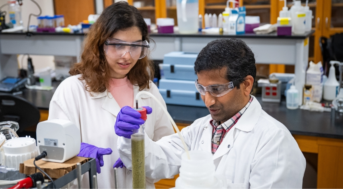 A faculty researcher and a student wearing lab coats, safety goggles and gloves examine a tall beaker of green liquid in a laboratory, surrounded by scientific equipment and bottles on lab benches.
