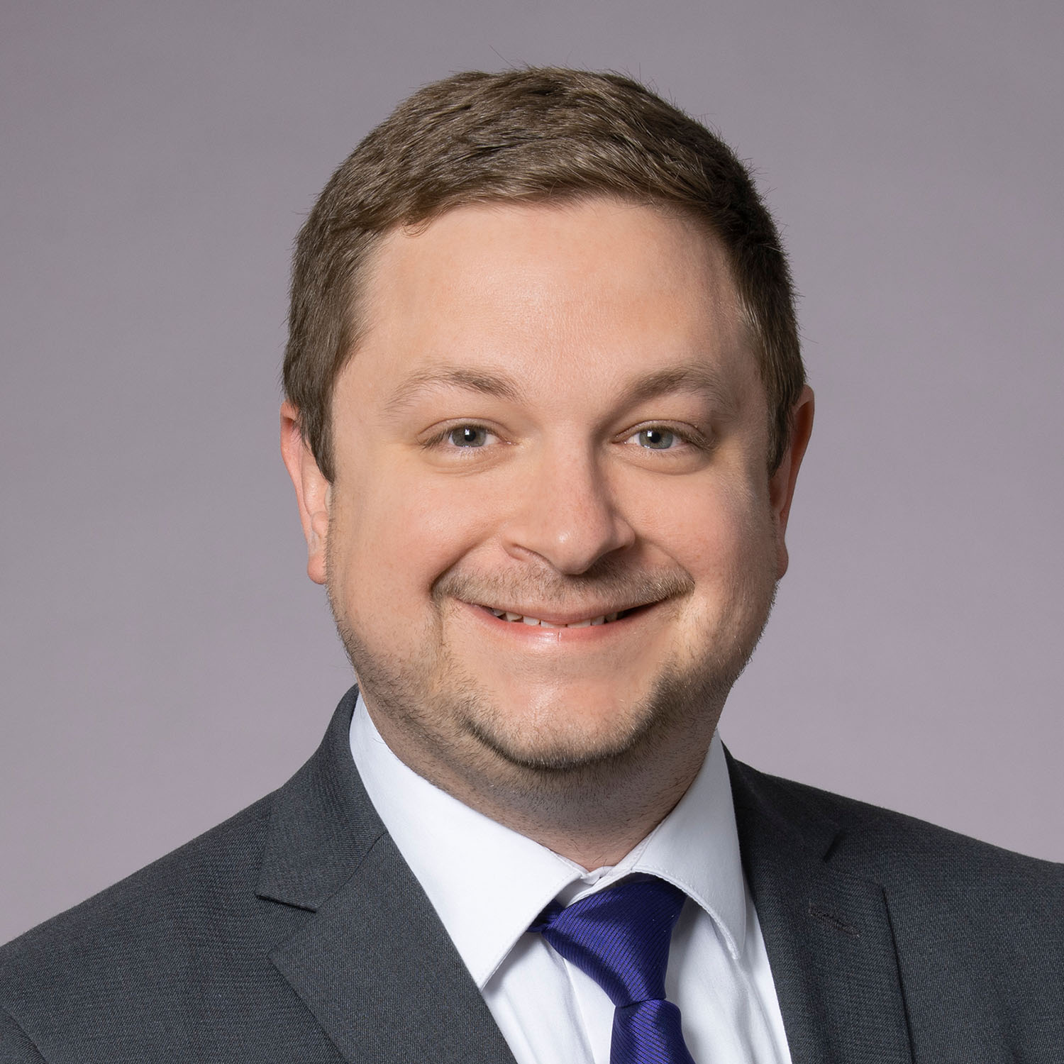 A man with short brown hair smiles for a portrait, wearing a dark grey blazer with a white collared shirt and a dark blue tie.