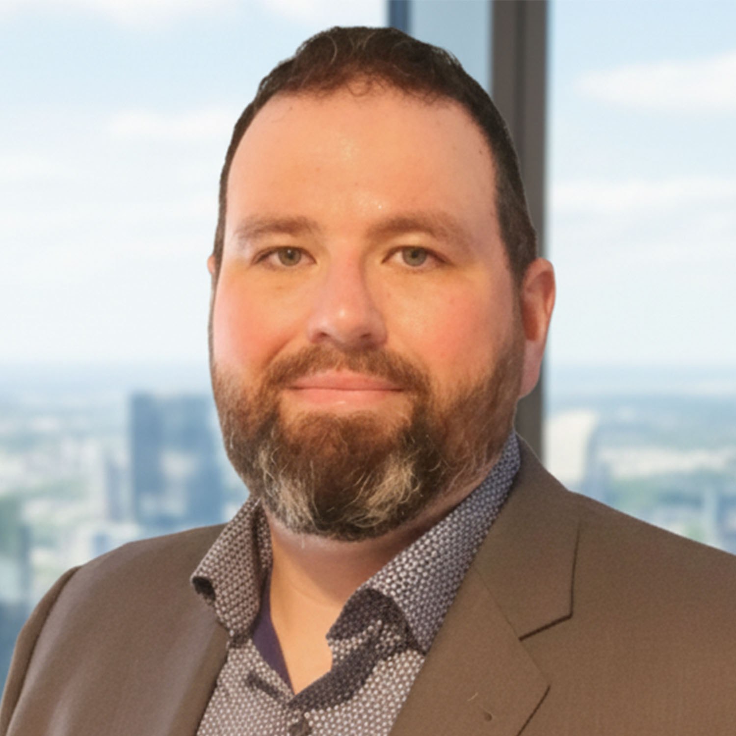 A man with brown hair and a beard and mustache wearing a gray blazer and a blue-and-white spotted shirt smiles against a window backdrop.