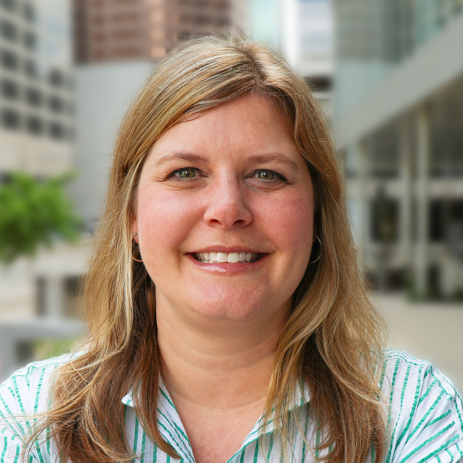 A woman with long blonde hair wearing a white and green-striped collared shirt smiles against an outdoor, urban, blurry background.