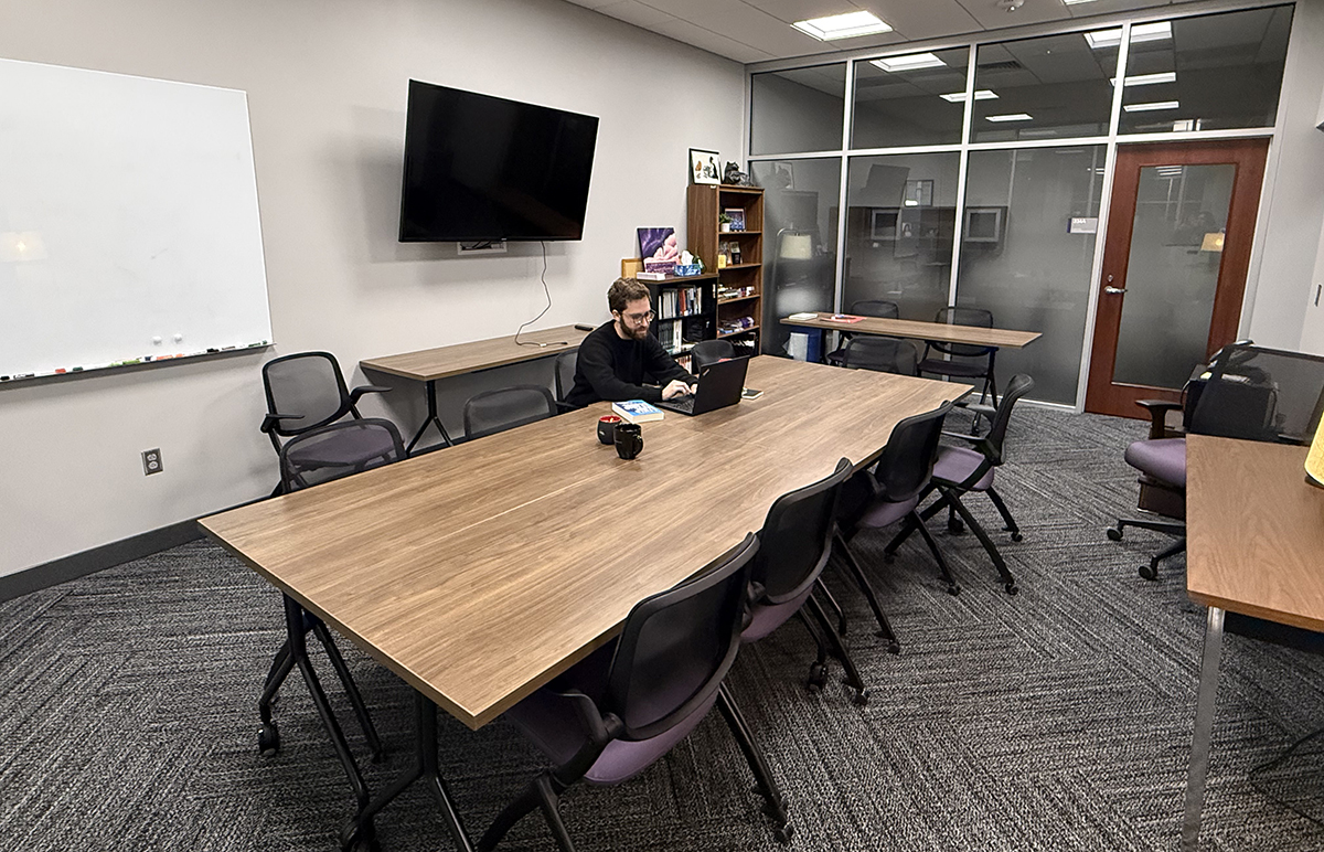 A man with brown hair and a beard works on a laptop at a table surrounded by purple chairs in a conference room.