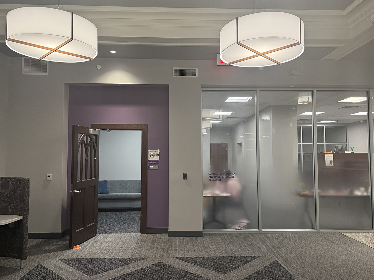 A doorway with a purple wall and a dark brown, open door is shown, with frosted glass windows showing into the room and the words "Scholar Development and Undergraduate Research" printed in purple.