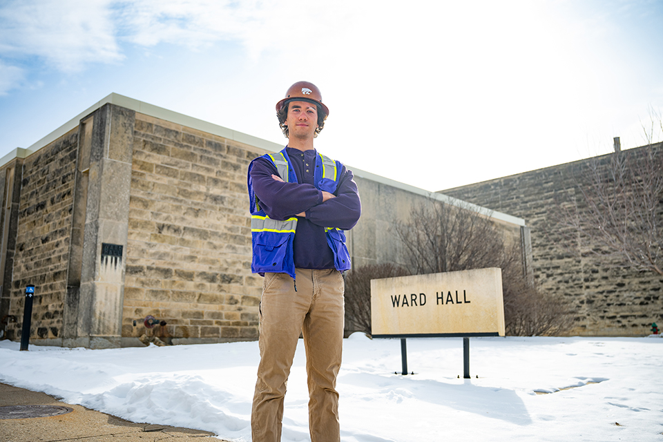 A young man wearing khaki pants, a purple long-sleeved shirt, a purple safety vest, and an orange hat stands in front of a limestone building near a sign that says Ward Hall.