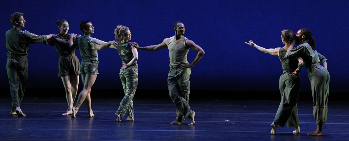 A group of dancers on a darkly lit stage with a blue background strike a dramatic pose in a line.