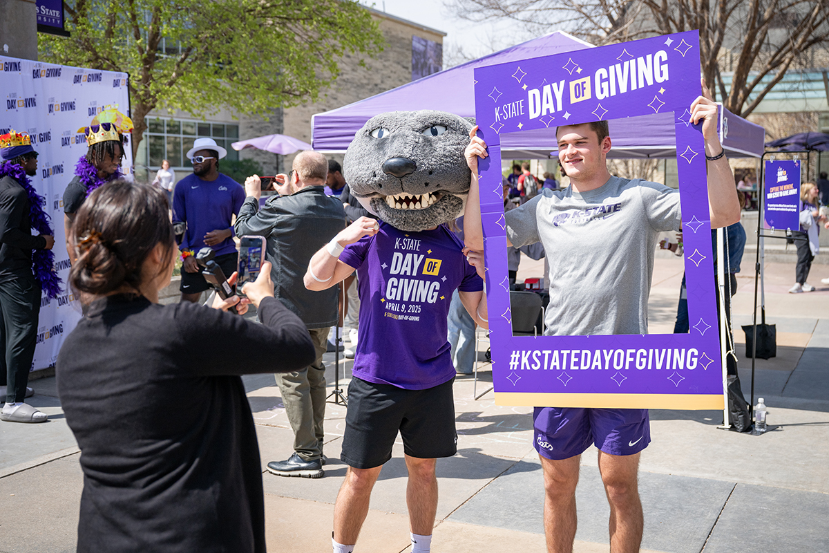 A student wearing purple shorts and a gray K-State t-shirt holds a K-State Day of Giving photo frame around his face while standing next to K-State mascot Willie the Wildcat.