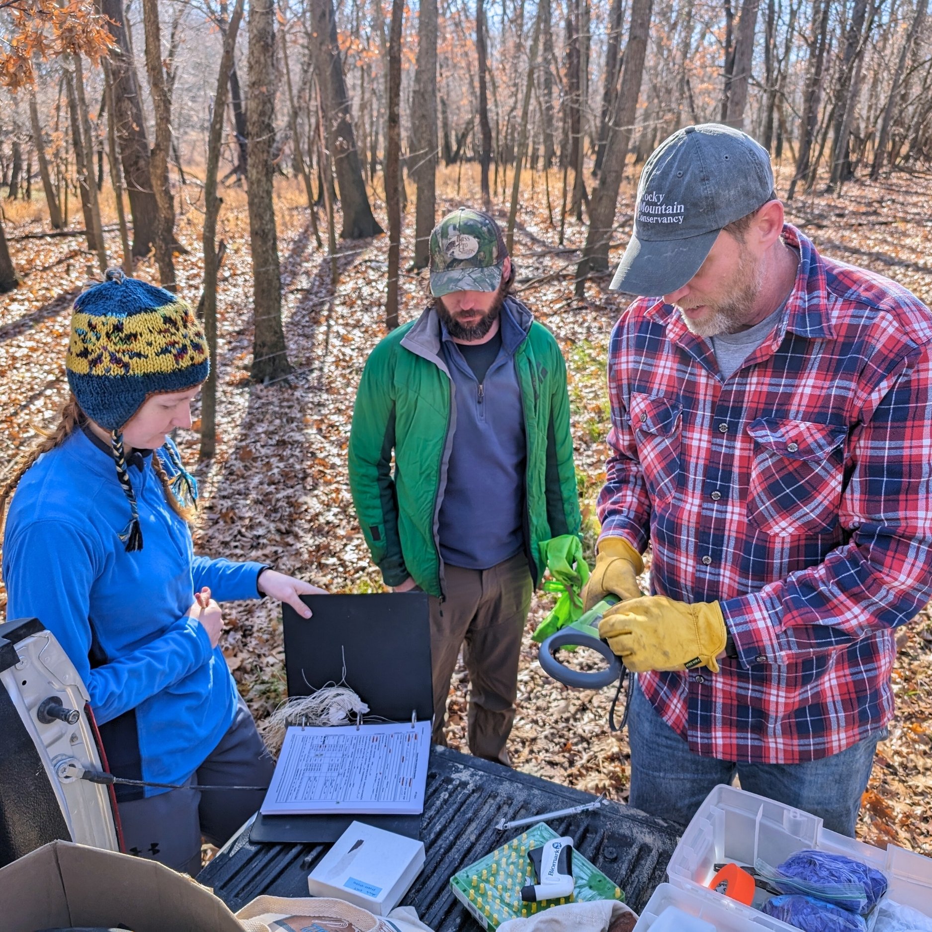 Adam Ahlers, wearing a red and blue plaid shirt and a Rocky Mountain Conservancy hat, explains field equipment to two students while preparing for a field study. Surrounding them is a fall forest, with barren trees and orange and red leaves.