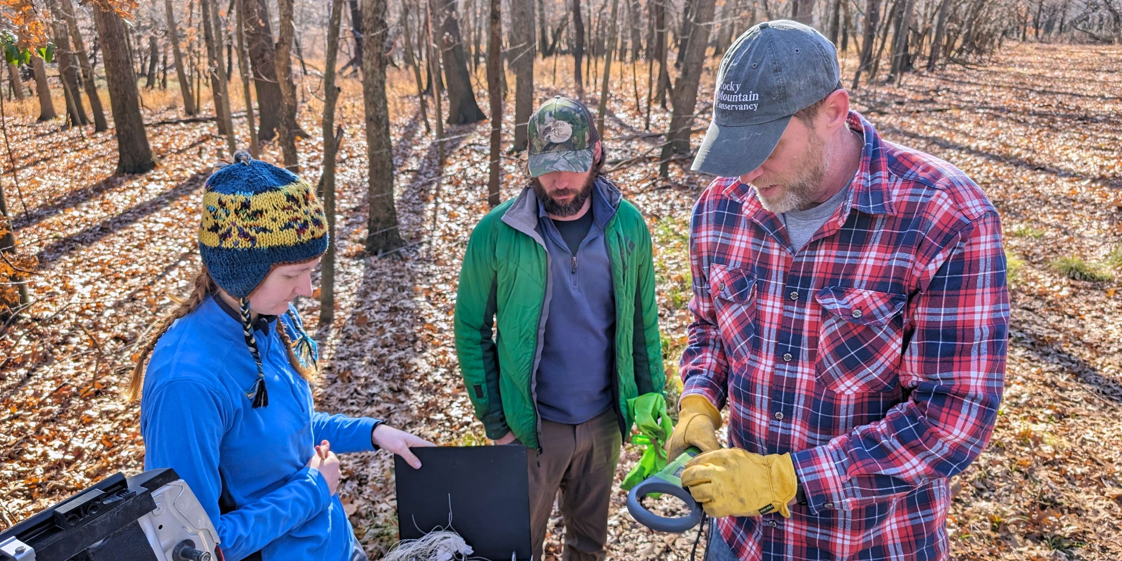 Adam Ahlers, wearing a red and blue plaid shirt and a Rocky Mountain Conservancy hat, explains field equipment to two students while preparing for a field study. Surrounding them is a fall forest, with barren trees and orange and red leaves.