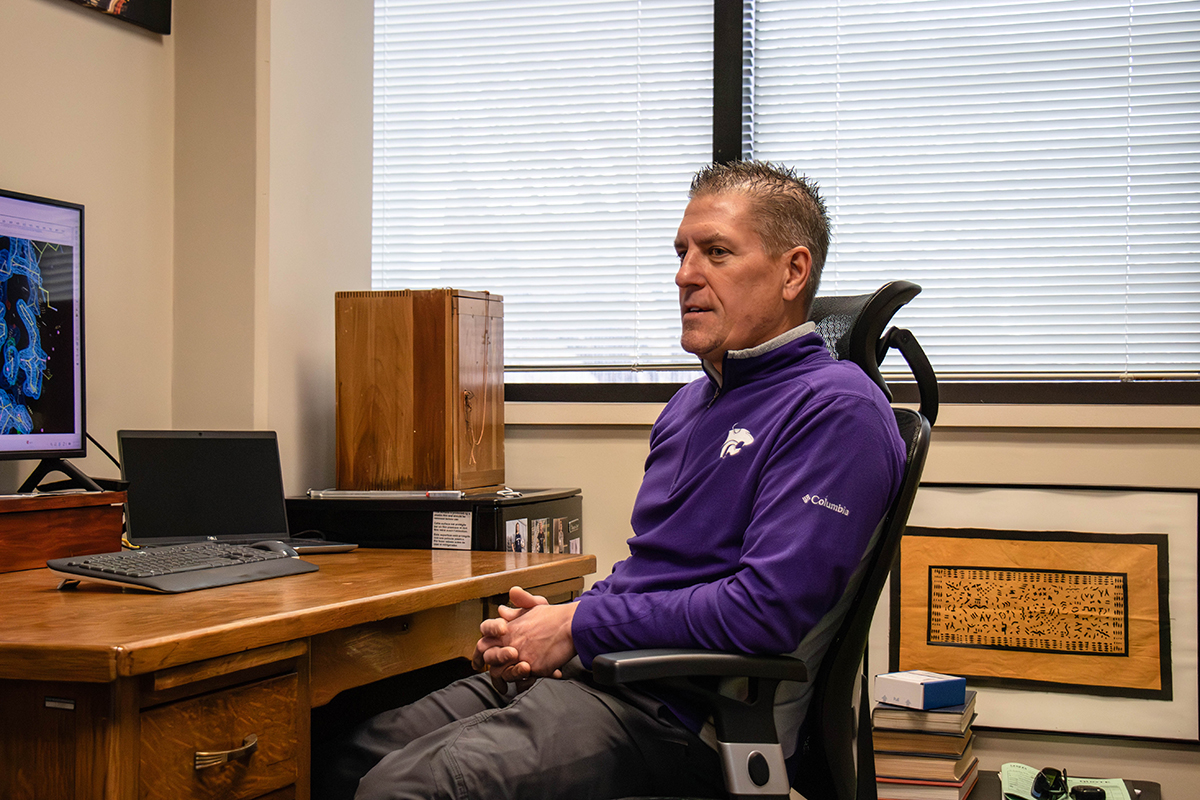 A male college professor in a purple fleece jacket sits at a wooden desk in his office. On his desk, a computer screen shows a colorful graphic of microscopic proteins.