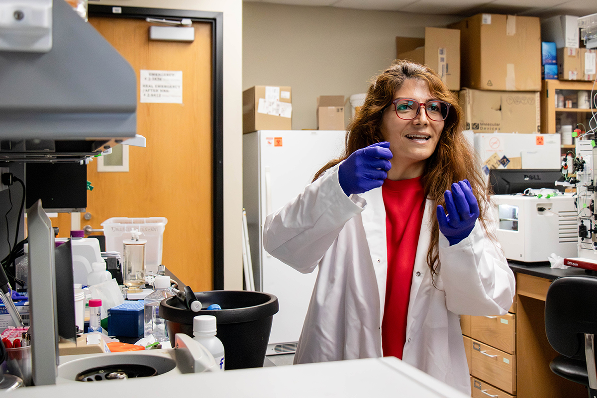 A female doctoral candidate in a white labcoat and blue gloves uses hand gesutres to explain a concept to the photographer inside a research laboratory.
