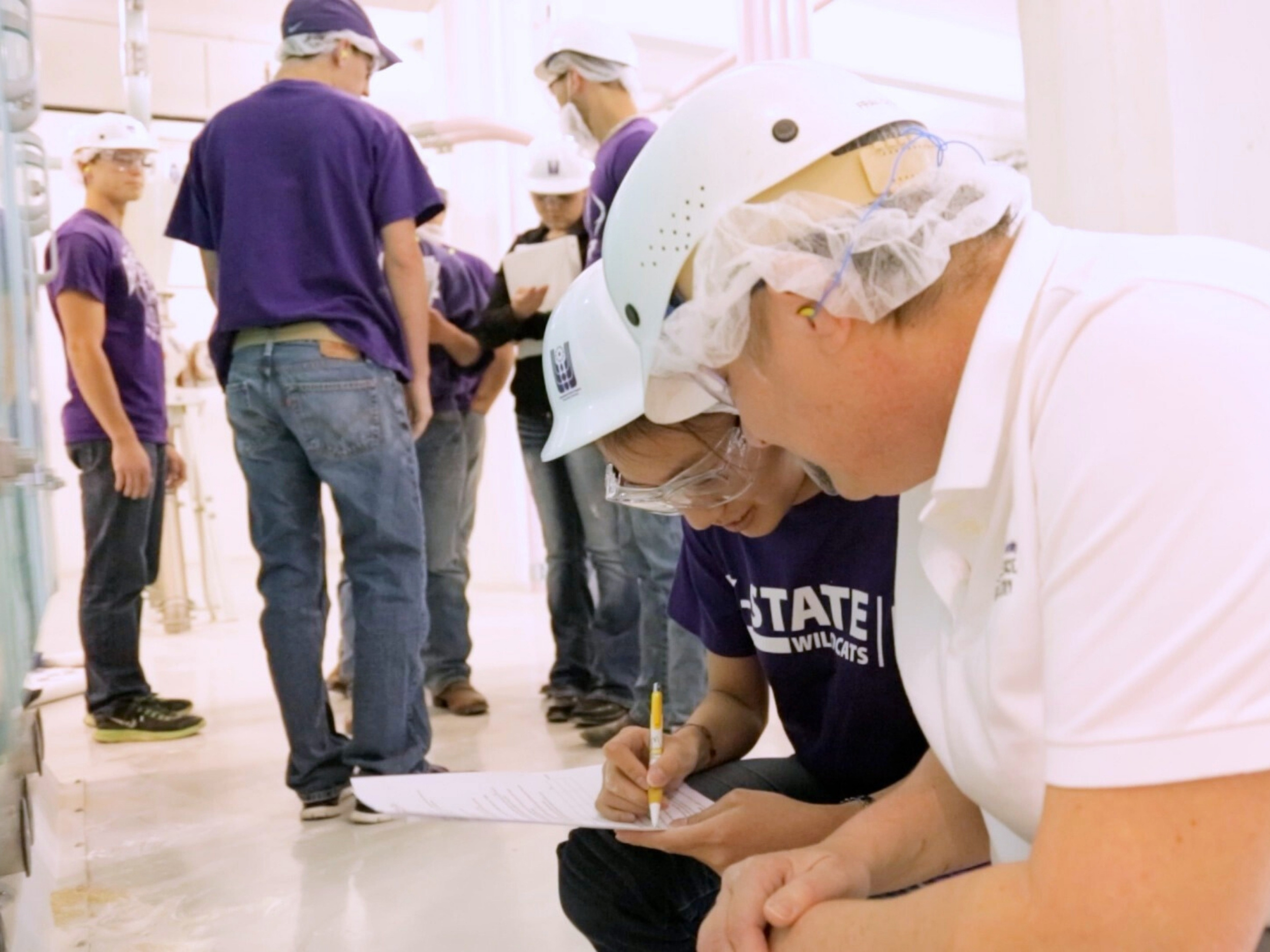 A female student and male teacher, both wearing white hardhats and goggles, crouch town to make note of research in the facility. The student, on the left, holds a yellow pen and writes down notes on a sheet of paper.