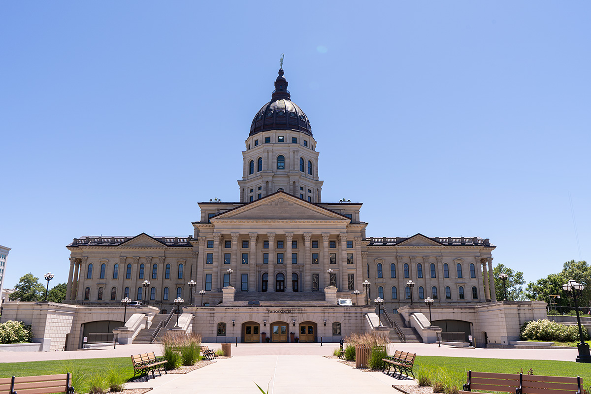 The Kansas Capitol, a rectangular biulding with a dark dome on top, stands against a blue sky with the walkway and front lawn leading up to the main entrance pictured.