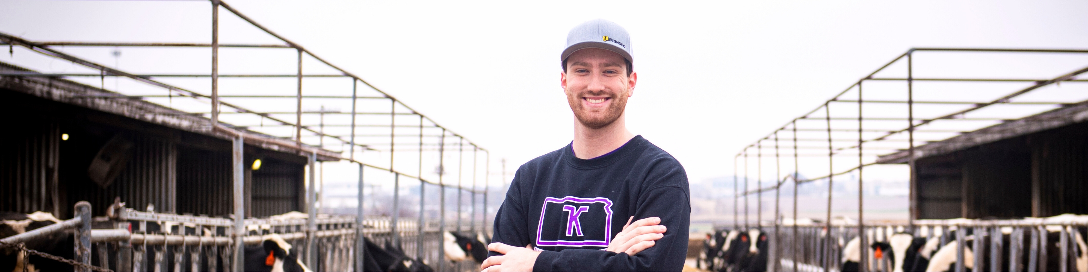 Carson Corwin, wearing a black sweatshirt and gray baseball cap, smiles with his arms crossed while standing in a feedlot aisle lined with Holstein cows in the background.