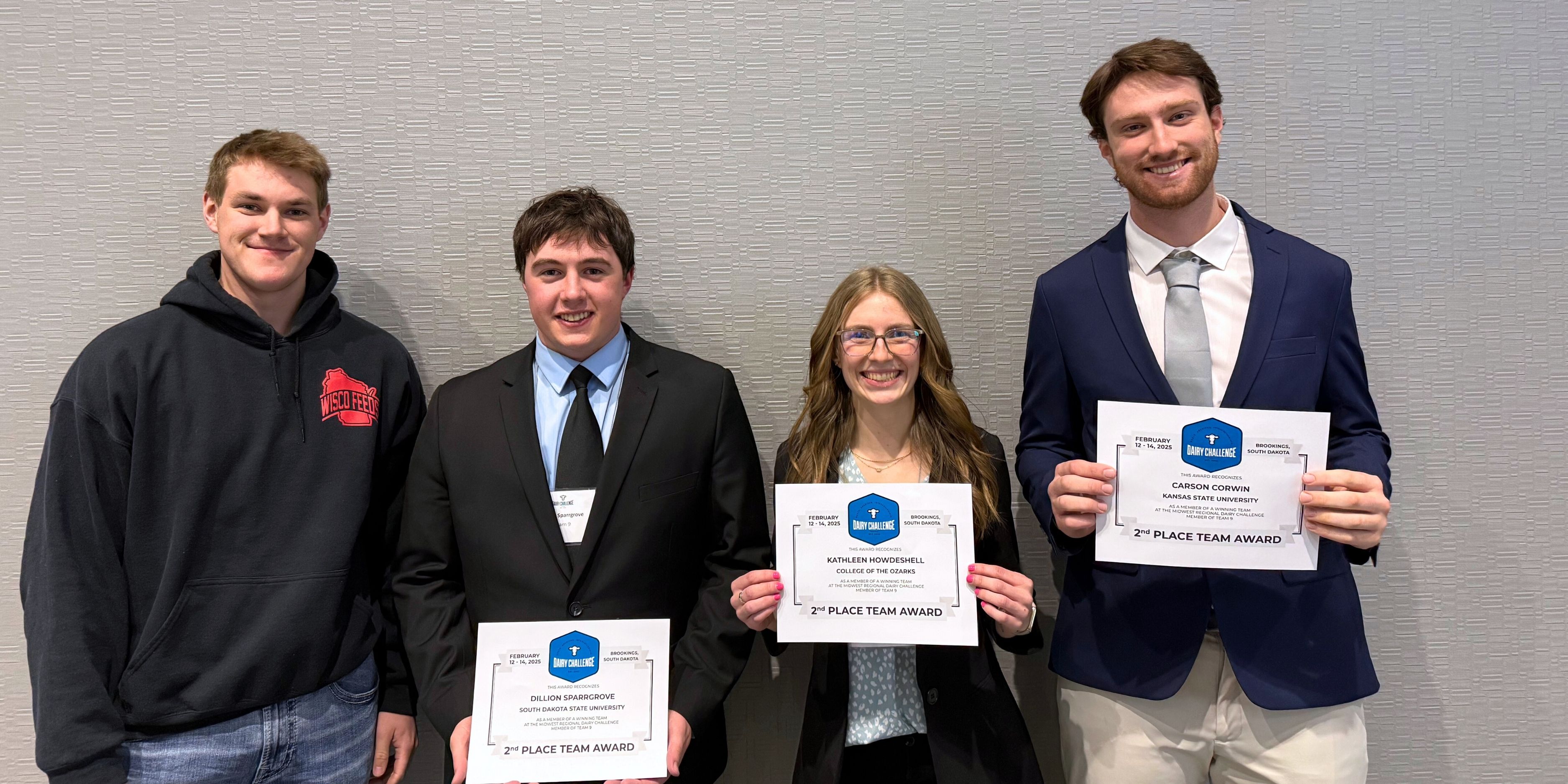 Four students stand in a line, with three holding certificates, at a Dairy Challenge competition. From left to right: a male in a black hoodie and jeans, a male in a black suit, a female in a black suit and Corwin, wearing a navy blazer and khakis.