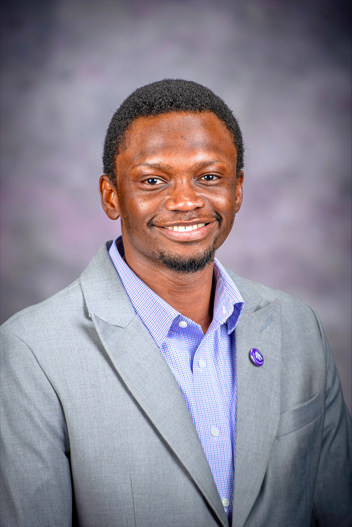 A graduate student wearing a light grey blazer over a purple shirt smiles against a dark grey backdrop.