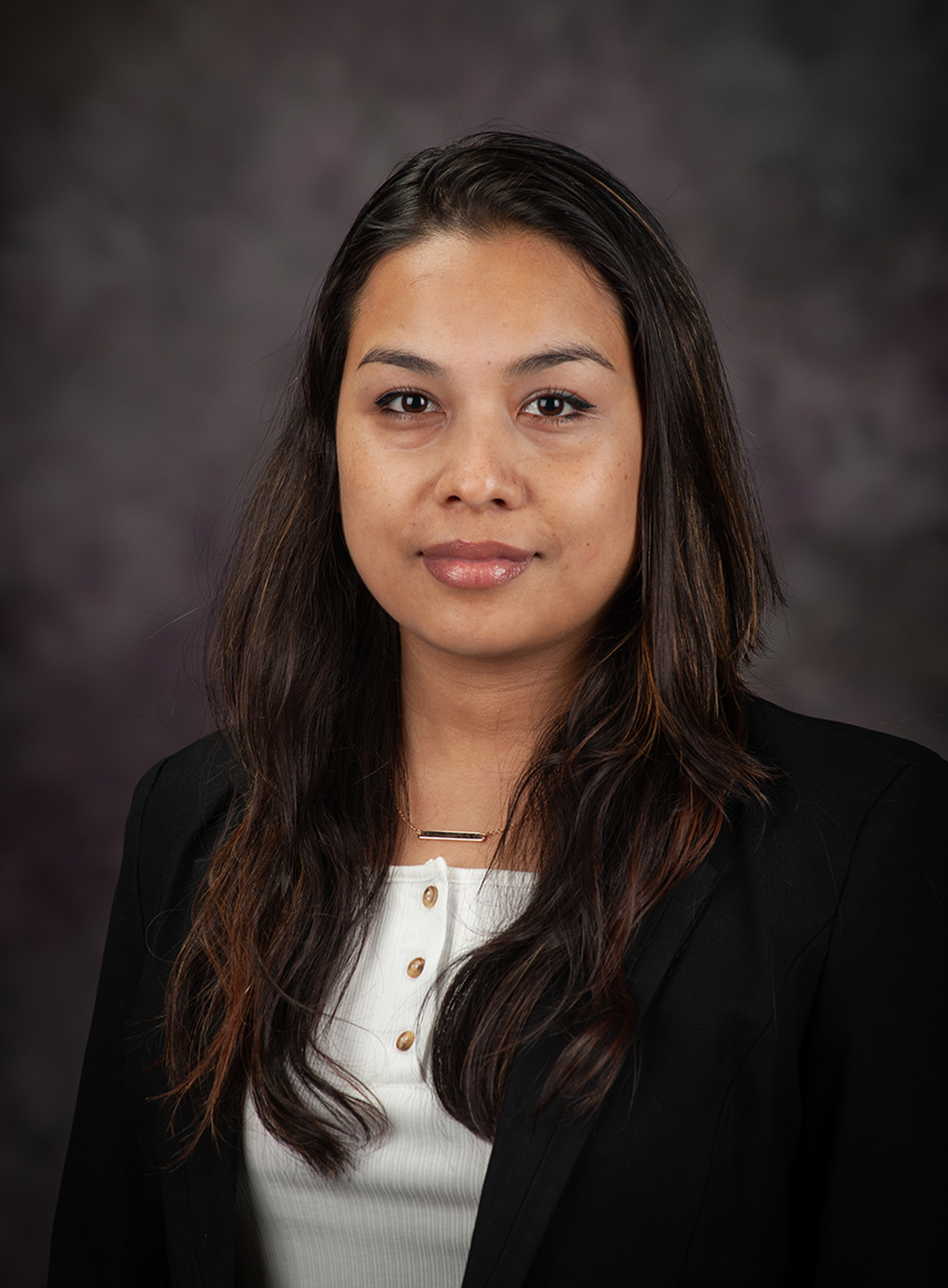 A graduate student with long dark hair wears a black blazer of a white shirt against a dark grey backdrop.