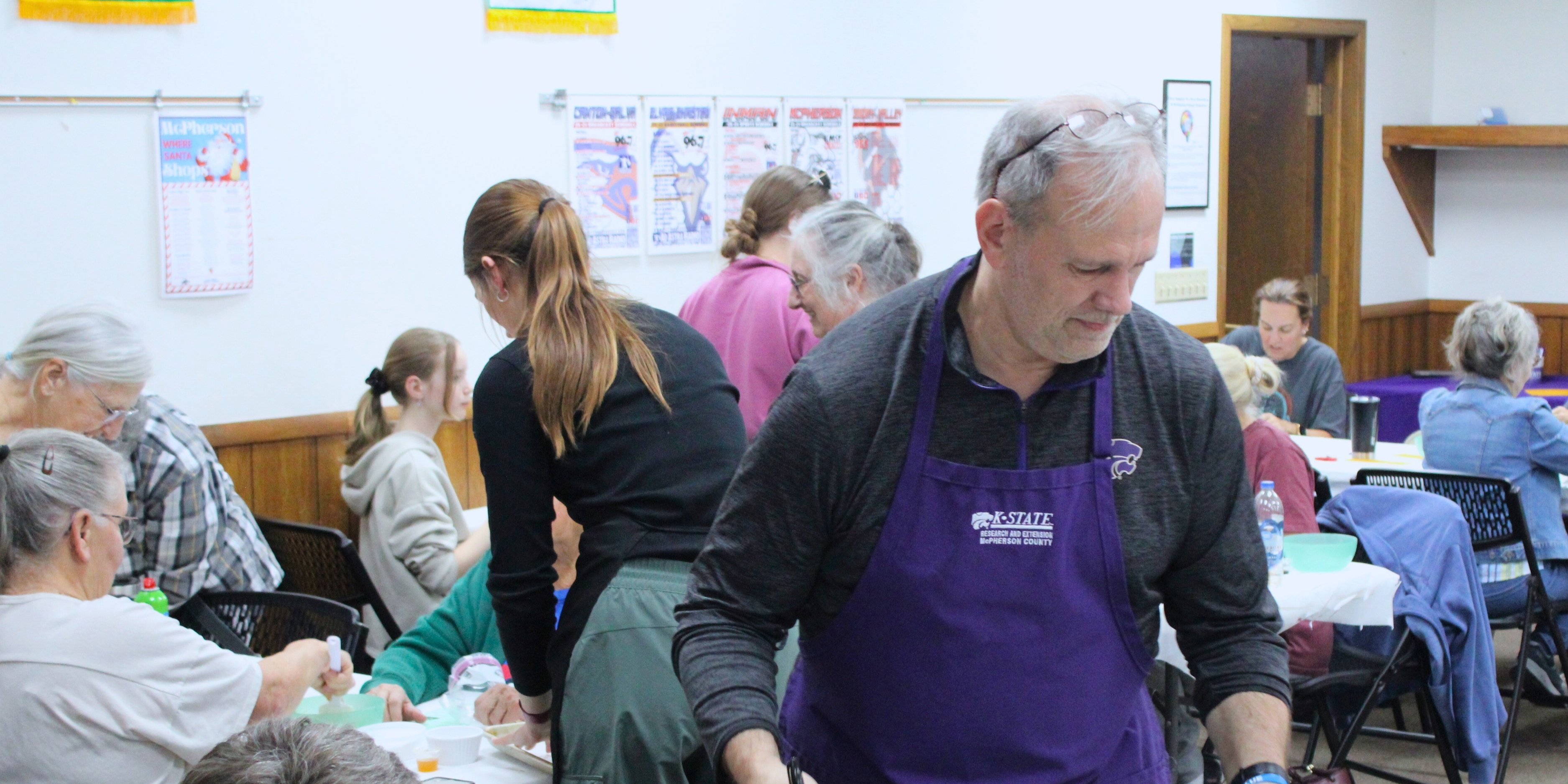 Chuck Laughlin, wearing a purple K-State Extension apron, offers supplies for a workshop on a baking sheet to participants. 