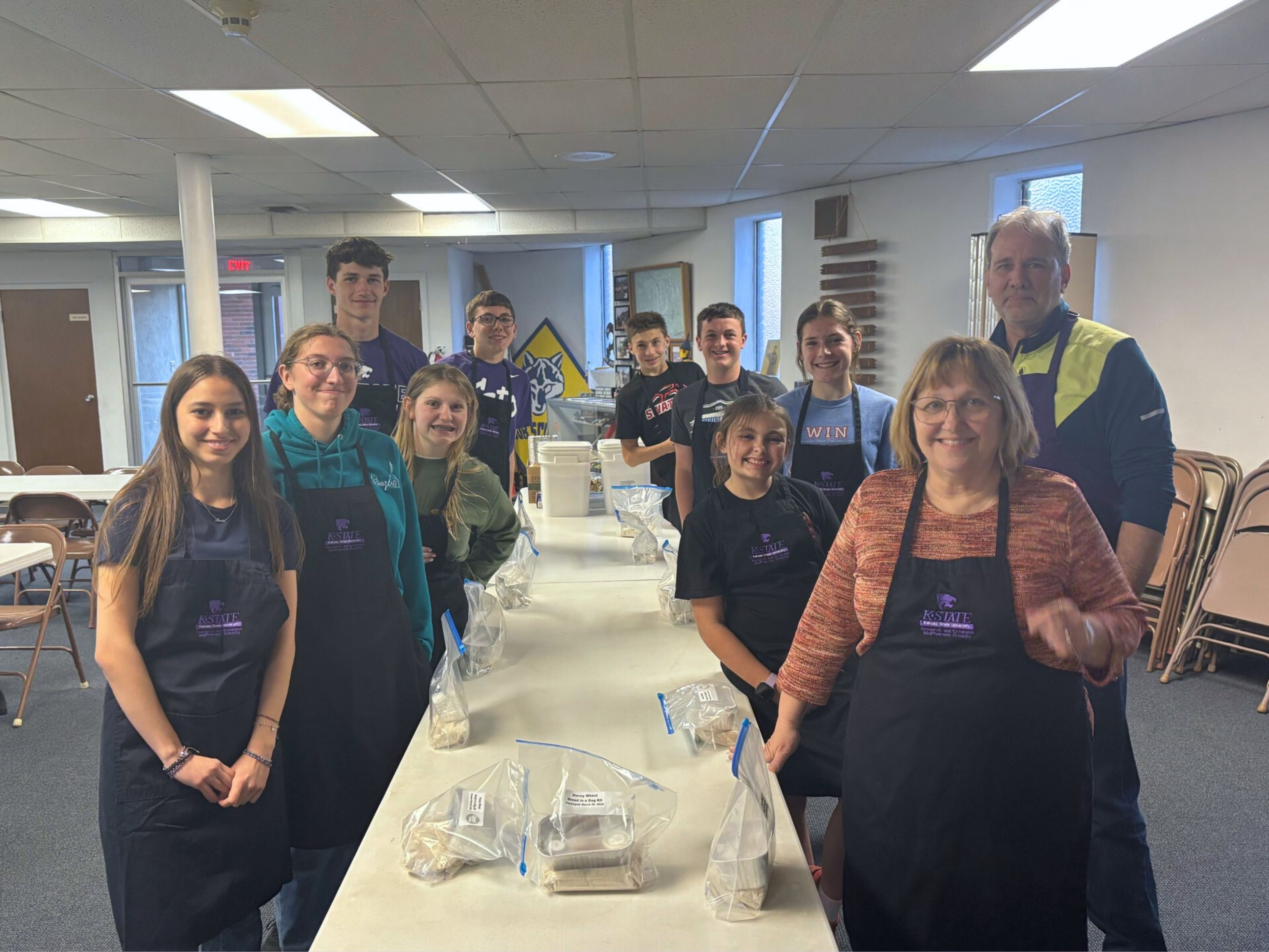 A group of smiling people in aprons stands around a table lined with plastic bags labeled "bread in a bag." They stand in a large, carpeted room with chairs lining the walls.