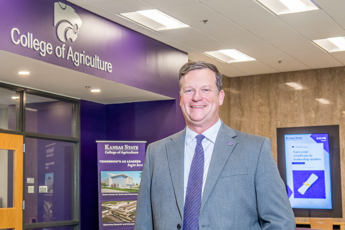 A male college dean in a gray suit and purple tie poses for a portrait in a college hallway outside of an office that has a purple powercat logo and the name "College of Agriculture" above the entrance.