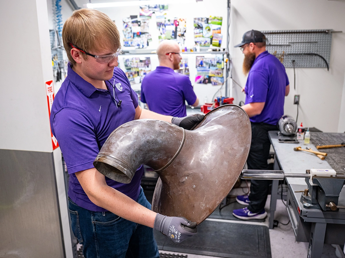 Holding the large, bell end of a rusted horn up against a tool, a male college student rotates the bell against the tool to smooth out small dents and imperfections in the instrument.