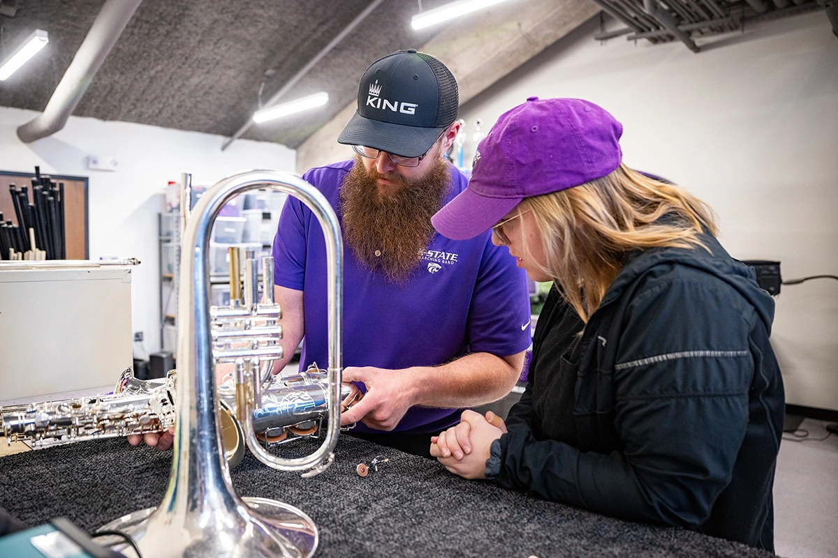A bearded male college instructor shows a female college student a spot on a saxophone they are working on at a musical instrument repair workbench.
