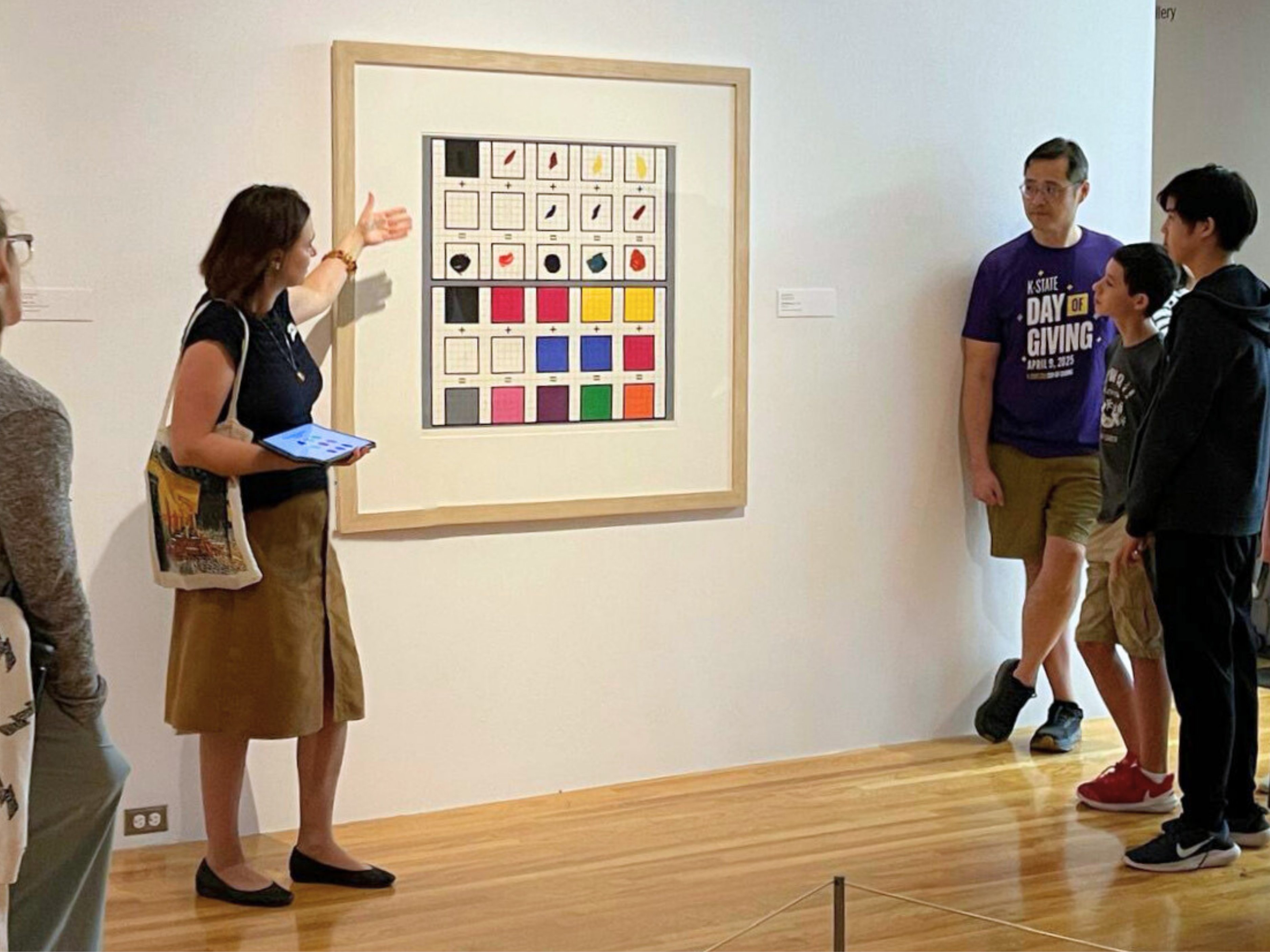 Natasha Rozhkovskaya presents to a tour group during a Beach Museum of Art event. She gestures toward a painting that features a white background with multicolor interlocking squares.