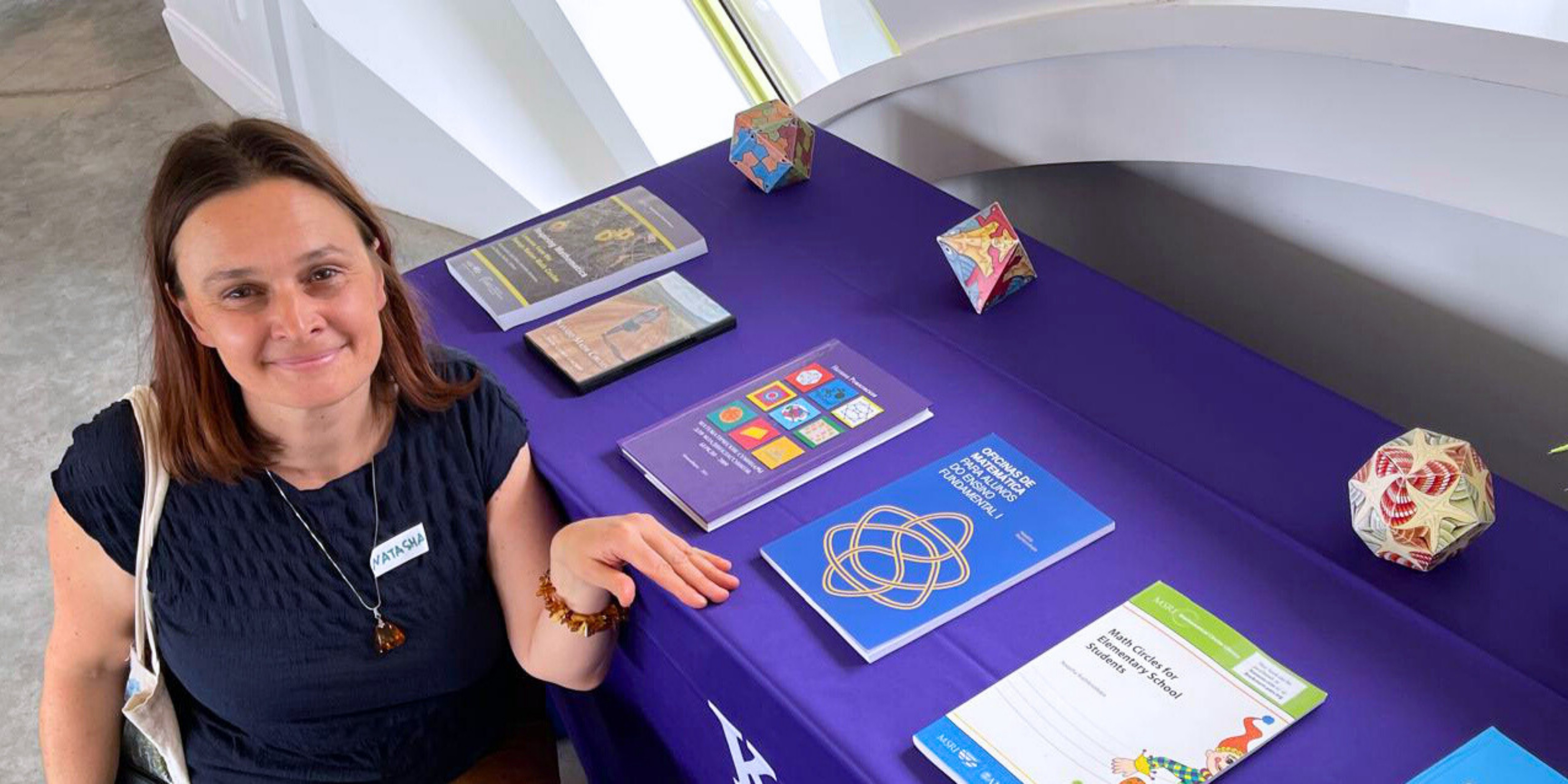 Natasha Rozhkovkaya, wearing a navy blouse, crouches next to a table displaying her authored books during a Beach Museum of Art event.