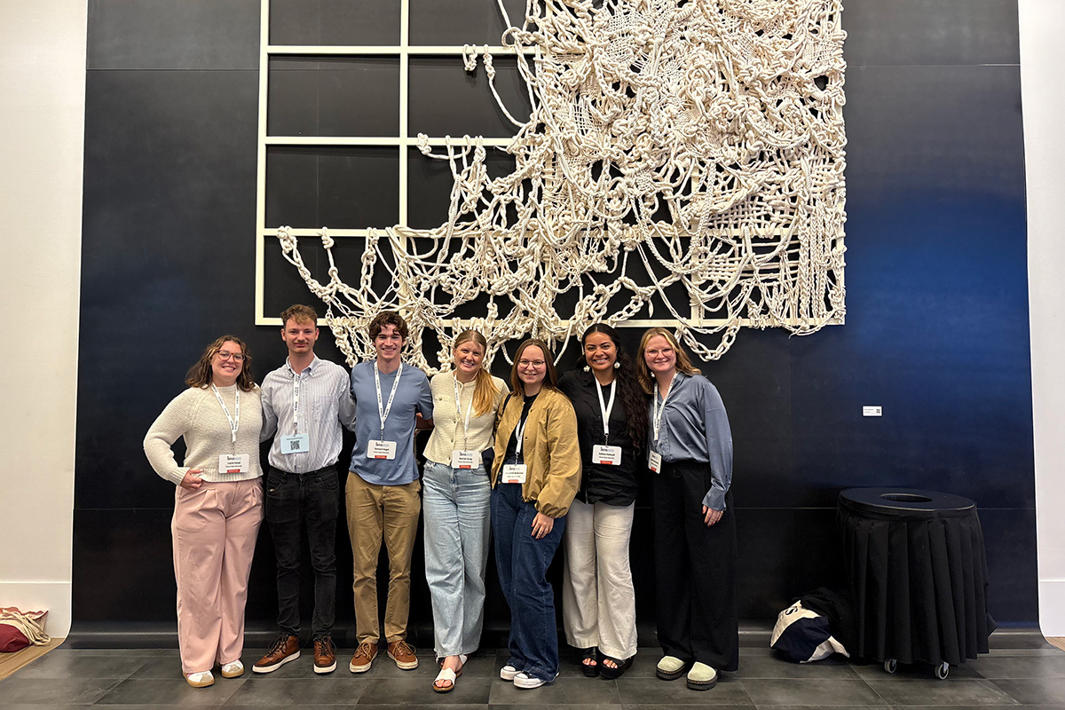 College students stand in front of a woven tapestry artwork against a dark wall. 