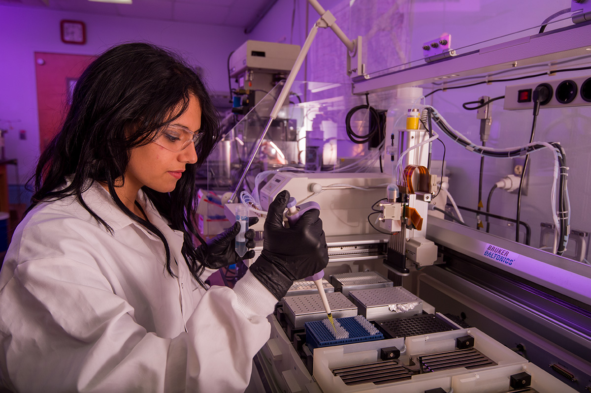 A scientist with long black hair wearing a white lab coat and black safety gloves dispenses materials into small tubes at a workstation in a lab with a purple hue.