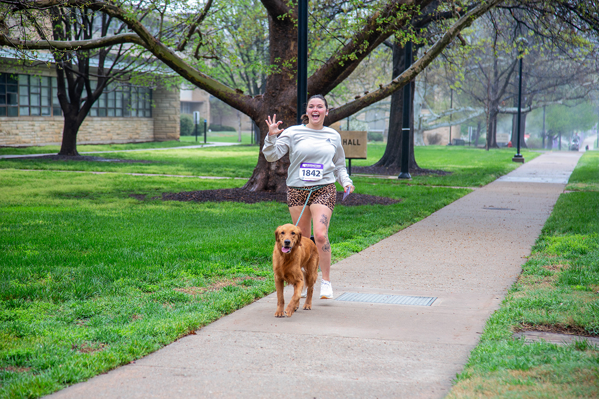 Smiling runner wearing white long-sleeve shirt, leopard print shorts and race bib 1842 waves at camera while jogging on campus sidewalk with golden retriever.