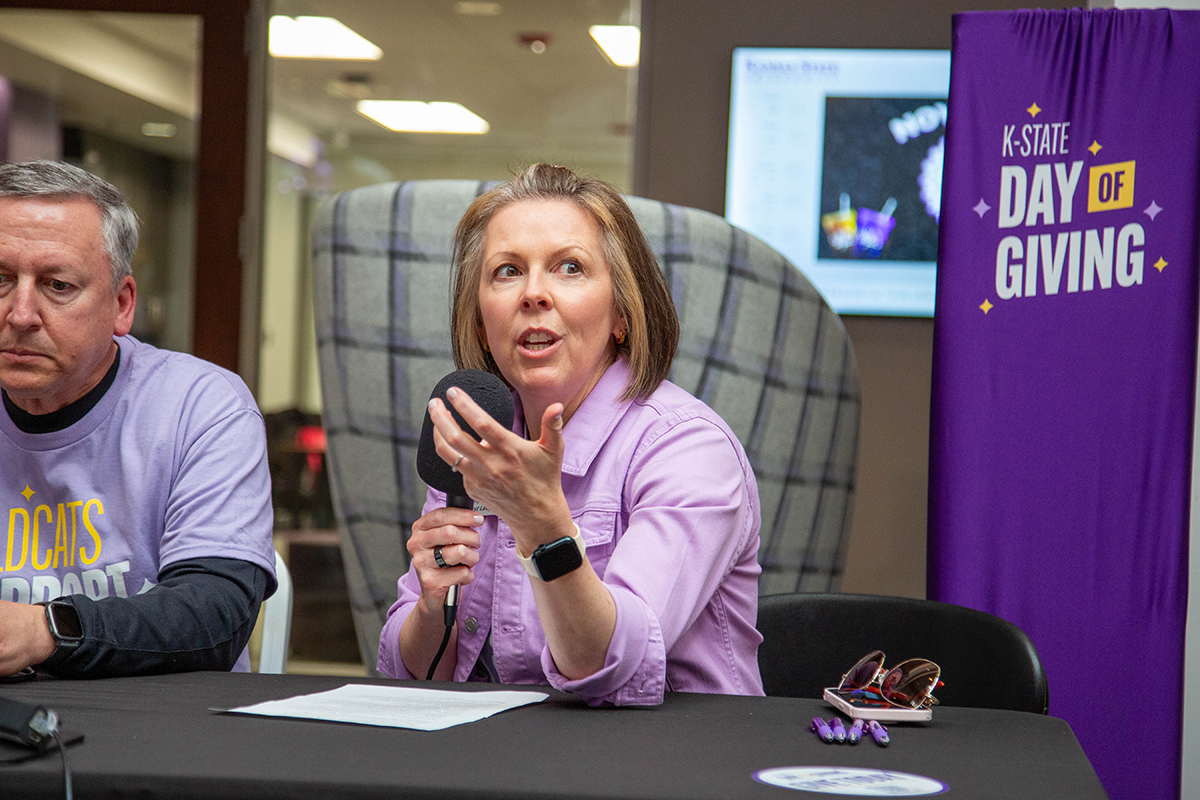 A woman in lavender shirt speaks into handheld microphone while seated at broadcast table with man in purple "Wildcats Support" shirt beside her. A Purple "K-State Day of Giving" banner stands to the right, with monitor and gray upholstered chairs visible in background. 