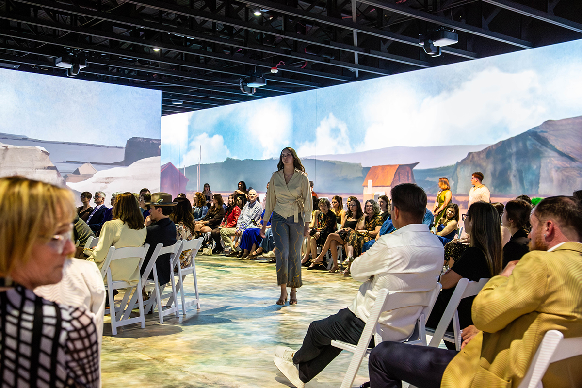 A model in beige wrap blouse and gray cropped pants walks down center runway toward camera wearing strappy heels. Audience seated on white folding chairs lines both sides of runway.