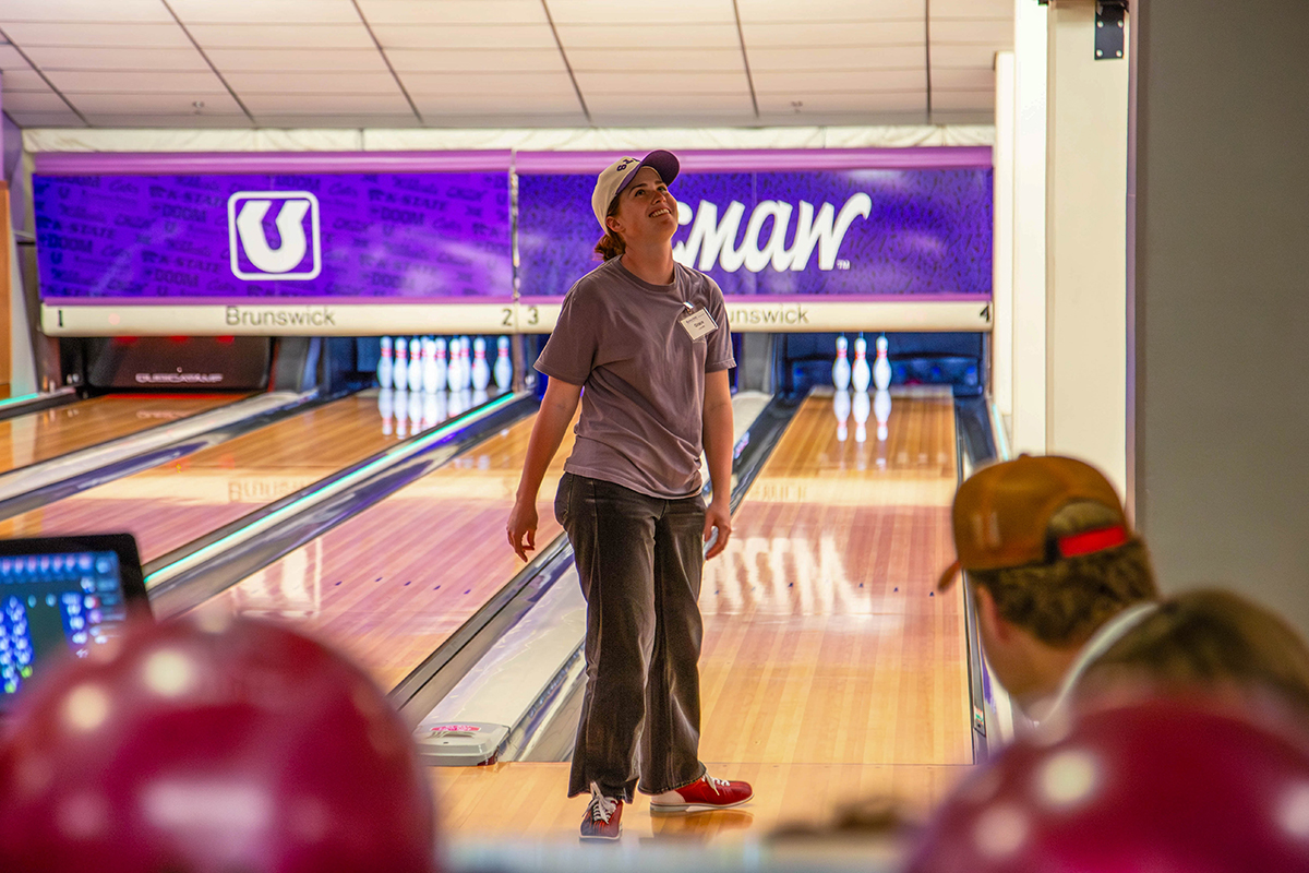 A person wearing tan baseball cap, gray t-shirt with name tag, and dark jeans stands on bowling lane looking upward, watching their ball.