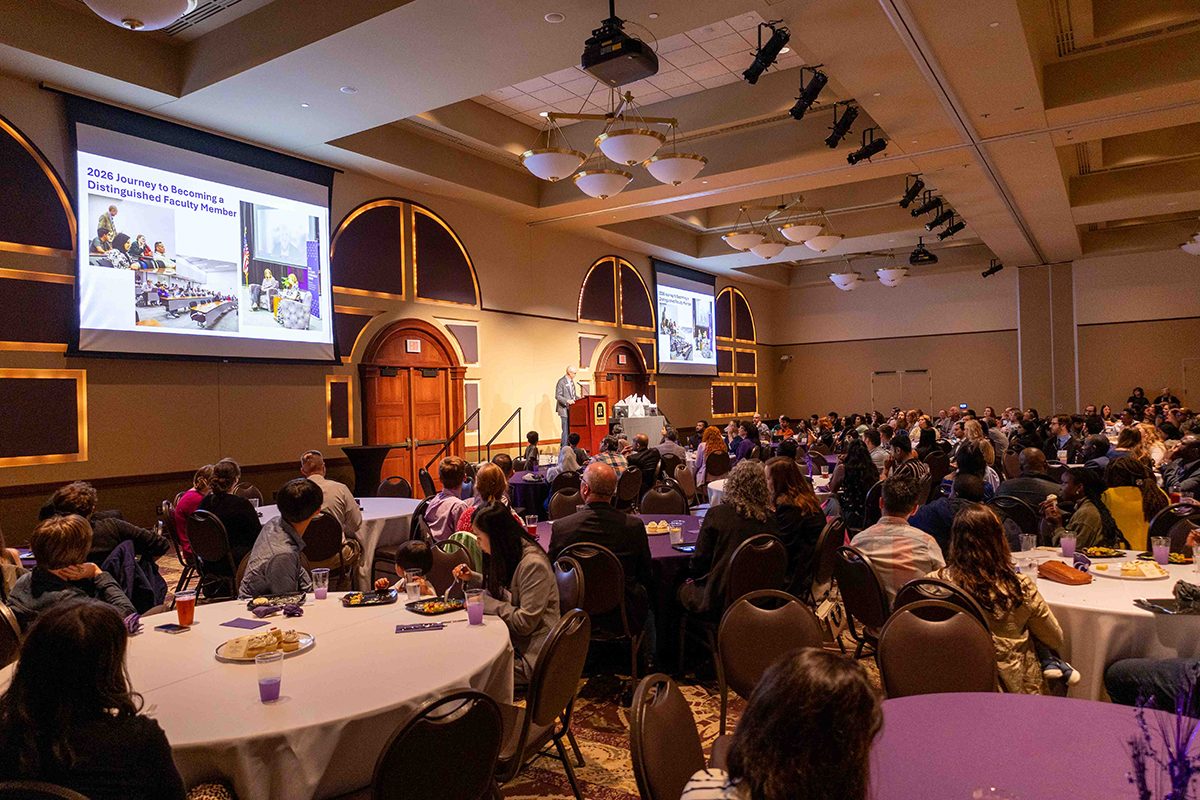  A crowded ballroom at Kansas State University filled with graduate students seated at round tables with purple and white tablecloths. A speaker stands at a podium on a stage with arched wooden doorways, while large screens display a presentation about becoming a distinguished faculty member. The room features elegant lighting fixtures and professional event staging.