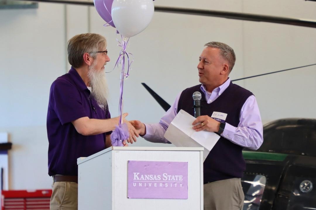Joel is on left wearing purple K-State polo shirt, khaki pants and glasses shakes hands with University President Richard H. Linton on right.