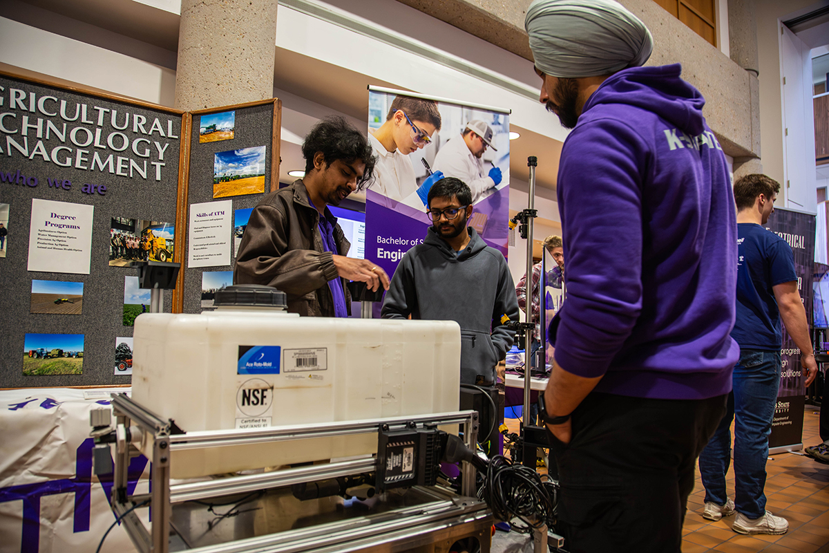Three students gather around white NSF-certified agricultural equipment with metal frame and mechanical components in foreground. Student on left wears black shirt with purple stole and gestures toward machinery while explaining. Student in center wears gray hoodie and glasses, listening attentively. Student on right wears purple K-State hoodie and gray turban. Display board behind them shows "Agricultural Technology Management" signage with photos of farm equipment, degree program information, and "Skills of ATM" section. 