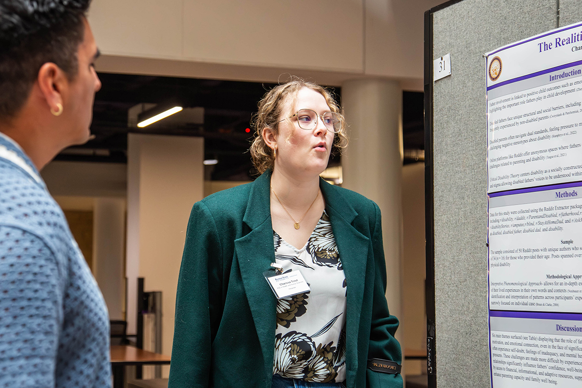 A master's student in a green blazer presents her research poster in a building courtyard.