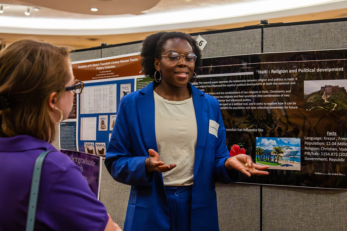 Student in blue cardigan and white shirt with name tag gestures while explaining research posters to attendees in purple shirt at conference. Poster titled "Haiti: Religion and Political development" displays research information about the topic. 