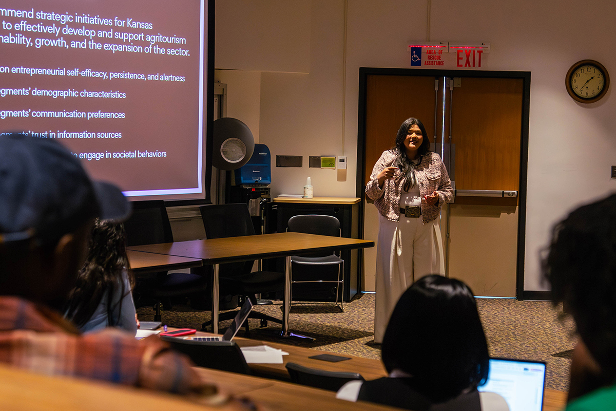 Presenter in patterned jacket and beige pants stands before seated audience delivering oral presentation. Large projection screen on the left displays a purple slide with white text about the research topic. 