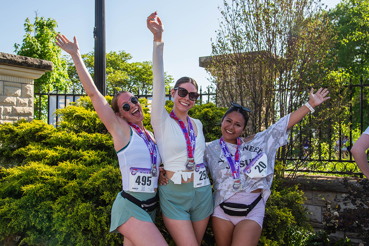 Three female runners wearing race bibs, finisher medals and purple beaded necklaces celebrate with raised arms and smiles in front of green landscaping and a stone pillar at the Run for the Roses 5K.