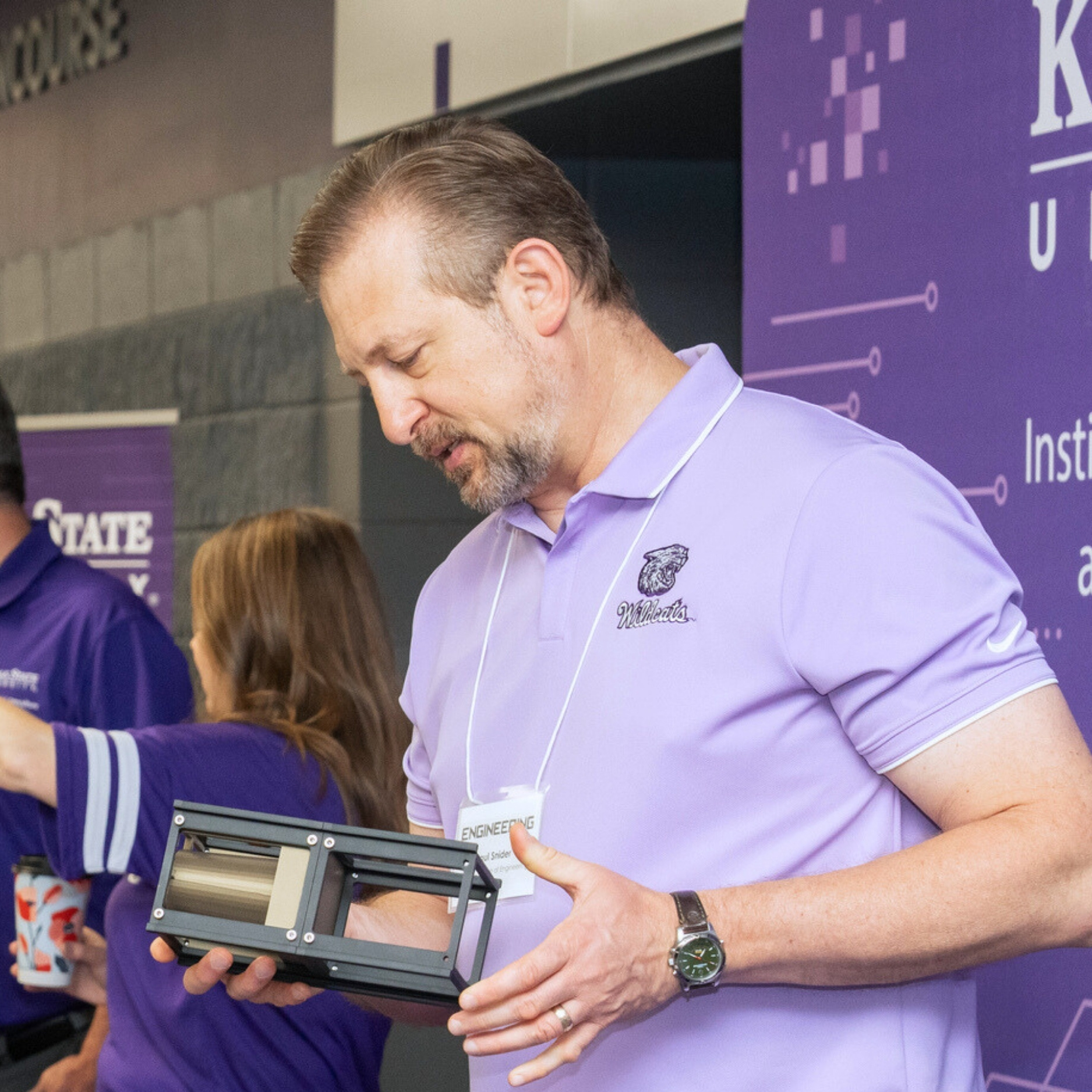 Paul Snider, in a lavender polo, stands at a table in Bramlage Coliseum for the Institute for Digital Agriculture and Advanced Analytics. Behind him is a purple pull-up banner. He speaks with two people: a man in a white polo and a woman wearing a blue crocheted headband.