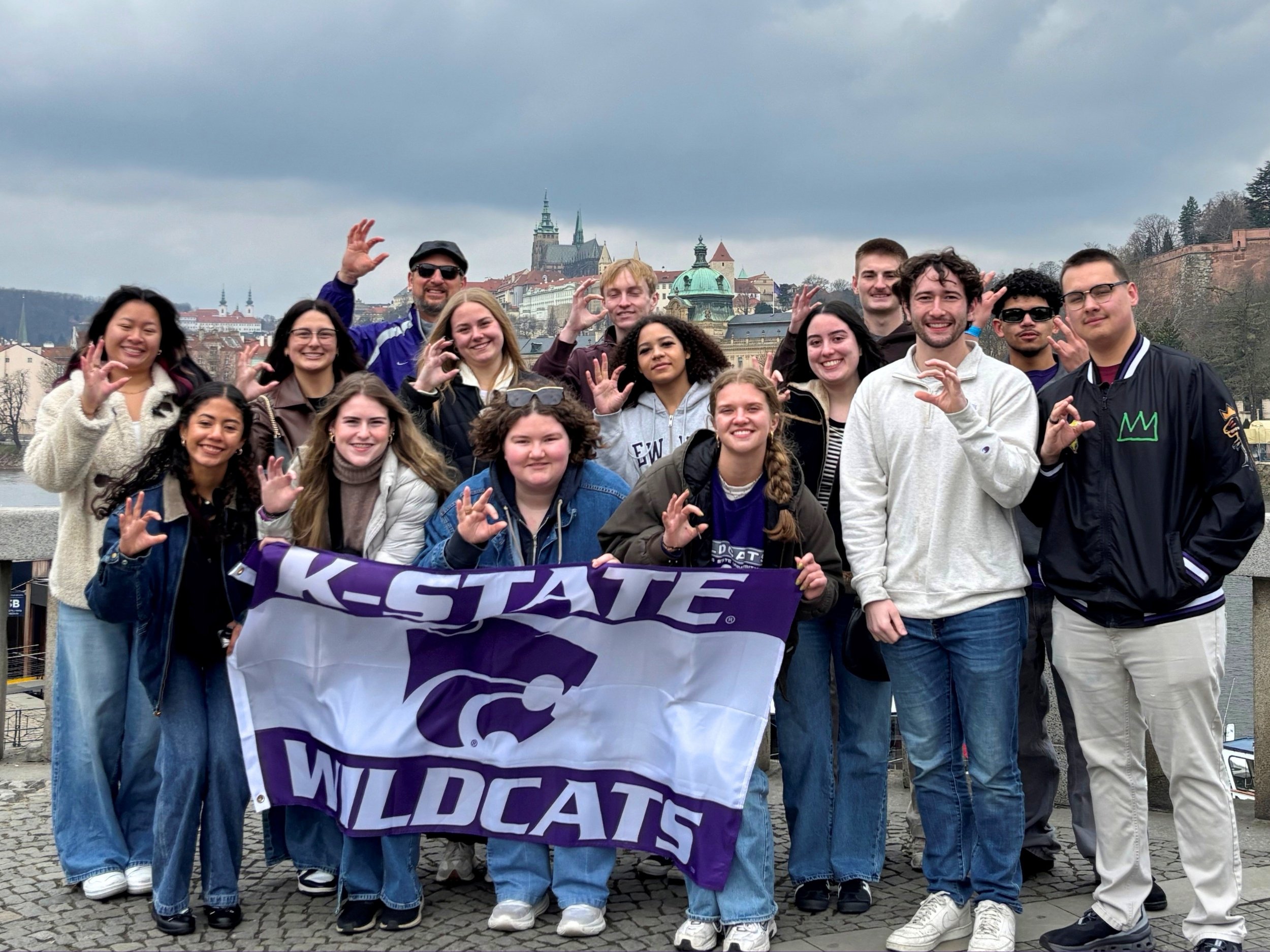 Snider and a group of students hold up powercat hand signs and a K-State Wildcats purple-and-white flag, smiling for a photo during their study abroad trip to Prague.