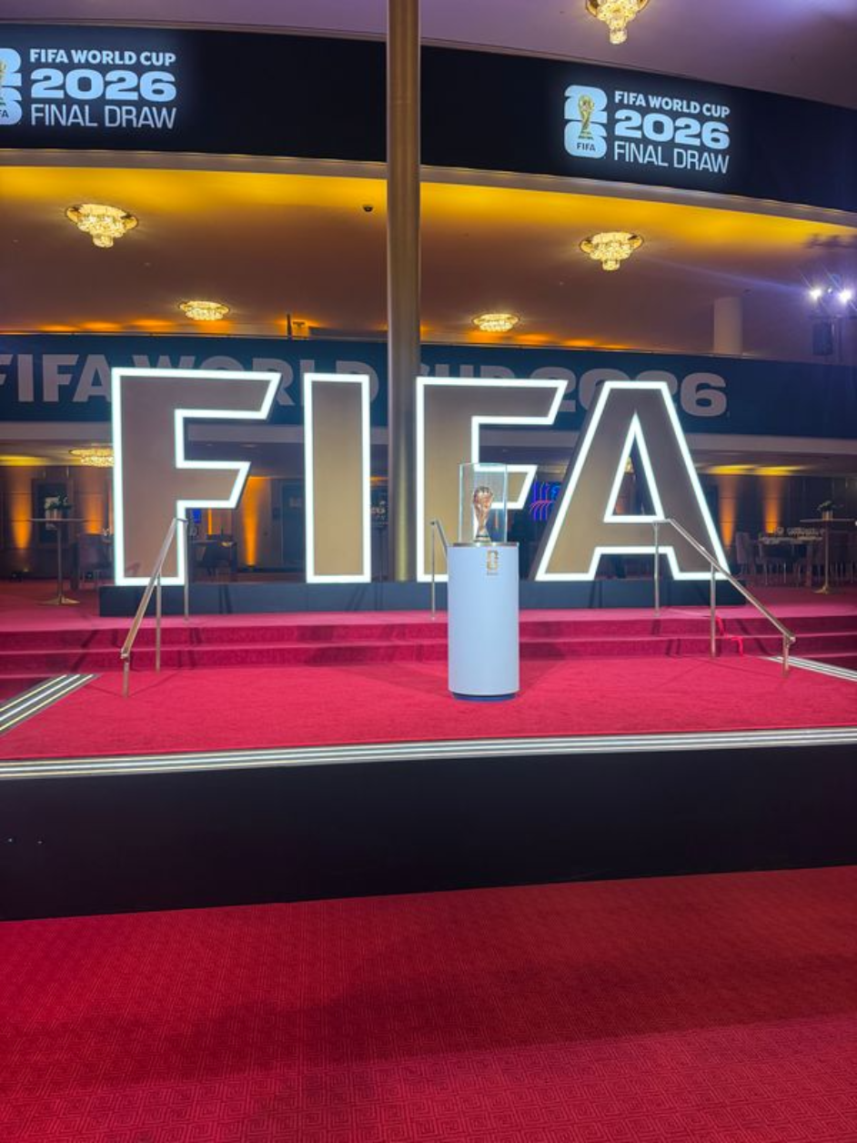 A red carpeted building has large letters spelling FIFA displayed behind a golden trophy on a pedestal.