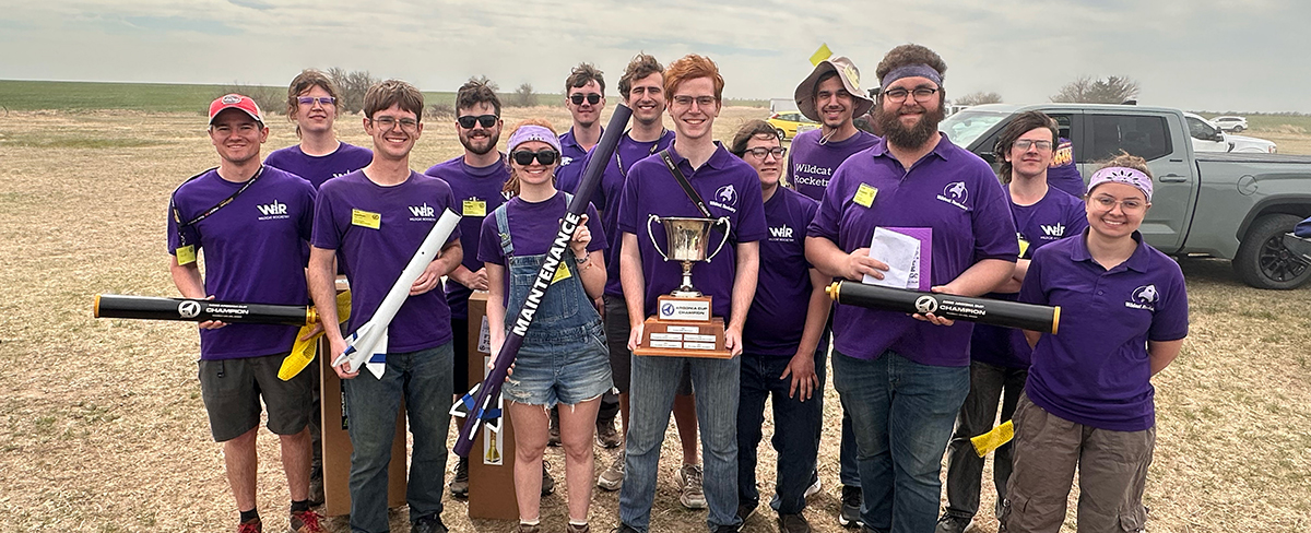 A group of students wearing purple polos and holding small rocket pieces and a large trophy smile as a group.