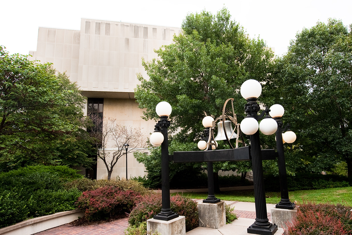 A large metal bell surrounded by iron lamps outside of a limestone building on Kansas State University's campus.