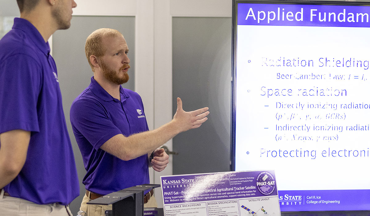 Two college students in purple polos give a presentation about the satellite they're building at a college open house.