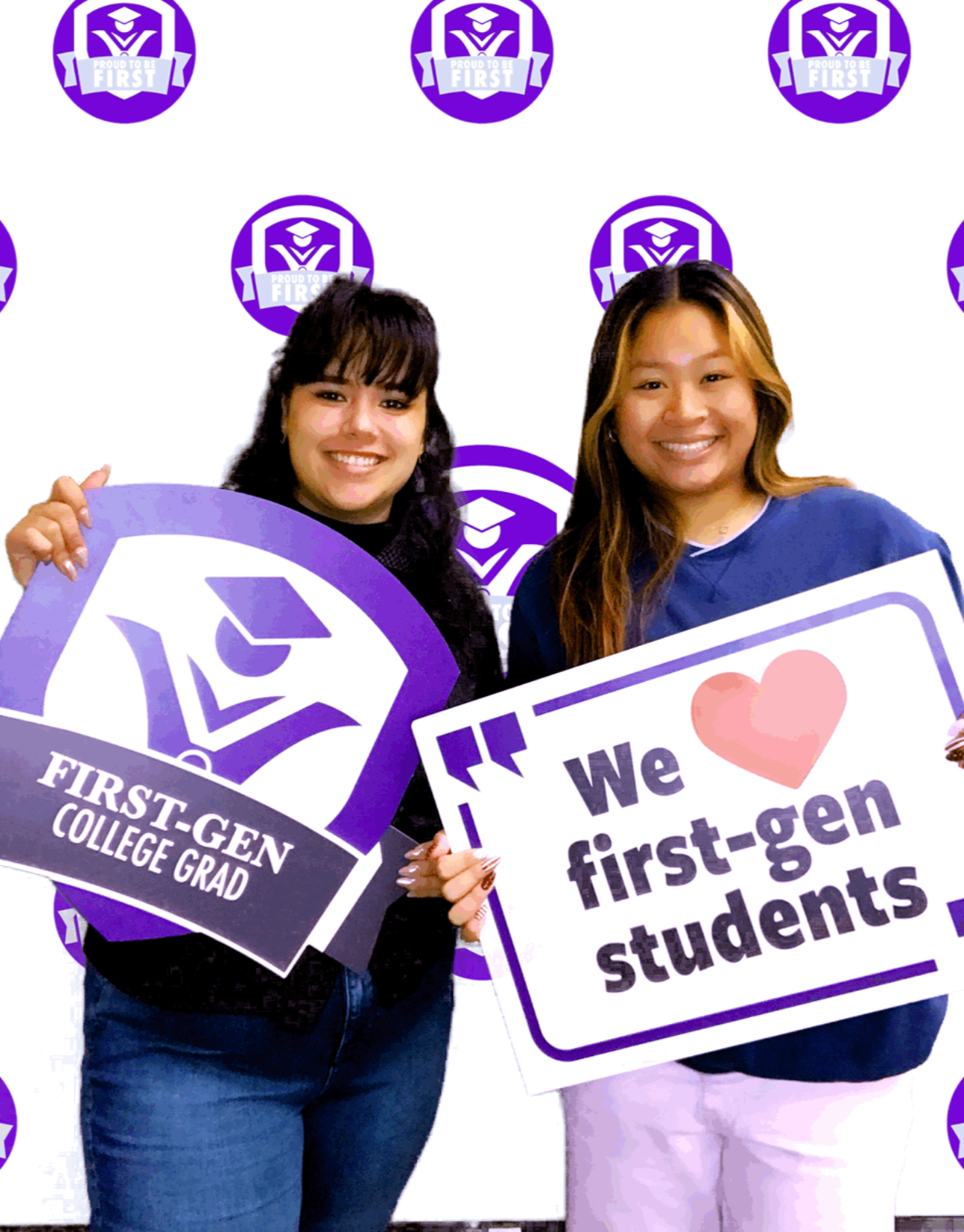 Two female students take a photo-booth-style photo holding first-gen student signs in front of a white background with purple circles that say proud to be first.