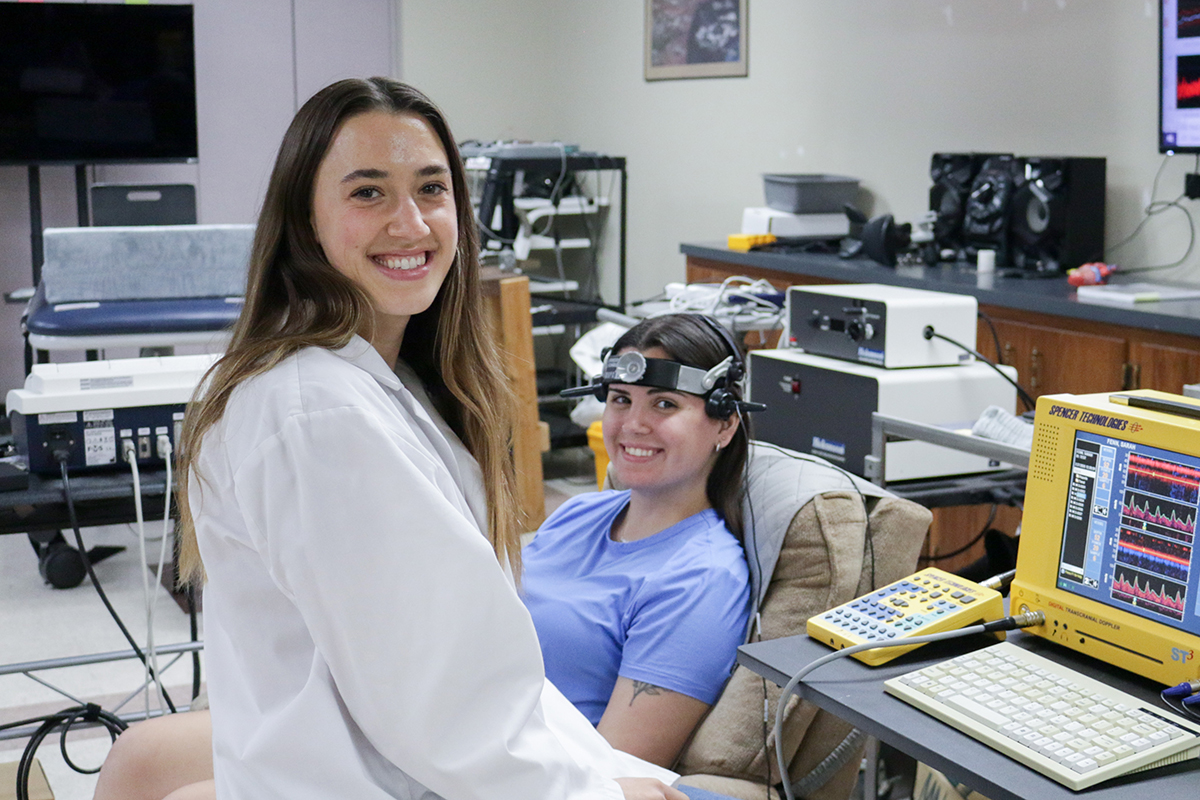 A female graduate student in a lab coat and a woman in an exam chair pose for a portrait inside an exercise lab.