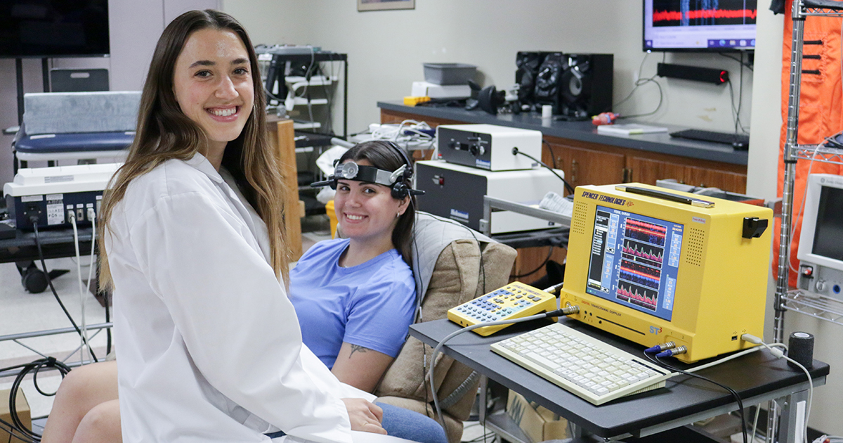 A female graduate student in a lab coat and a woman in an exam chair pose for a portrait inside an exercise lab.